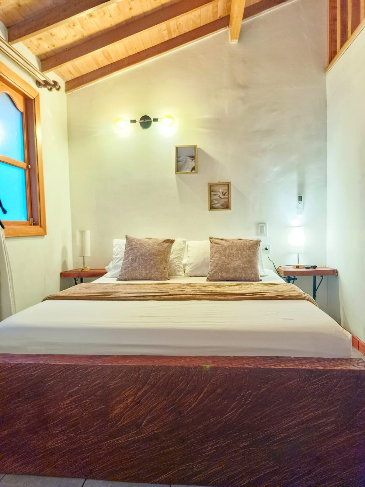 A bedroom with a wooden ceiling, a bed with white bedding and two pillows, a bedside table with a lamp, and a wall-mounted light fixture.