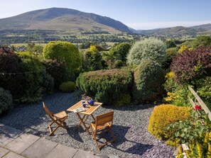 A garden with a table and chairs, surrounded by various shrubs and plants, with a mountainous landscape in the background.