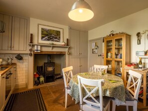 Una cocina acogedora con estufa de leña, armarios blancos, una mesa de comedor con sillas y una estantería.