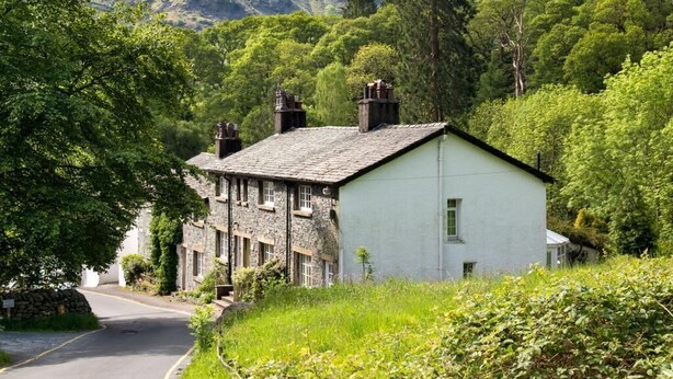 A stone house with a white exterior, surrounded by lush greenery and mountains in the background. - 2 Bed in Keswick (oc-sz068) (Borrowdale)