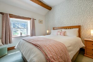 Ein Schlafzimmer mit einem großen Bett, einem Holz-Kopfteil, einem Fenster mit Vorhängen, einem Nachttisch mit Lampe und einer Wand mit Muster-Tapete.