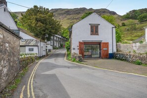 A white building with a brown door and window, a stone wall on the left, and a road with double yellow lines. - 3 Bed in Borrowdale (oc-sz465) (Borrowdale)