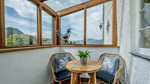 Uma sala de sol com vista para as montanhas, uma mesa de madeira, duas cadeiras de vime e um vaso com planta.