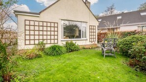 Una casa a un piano con tetto spiovente, un camino a vista e un piccolo giardino anteriore con prato e una sedia in metallo.