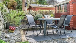A patio with a table and chairs, surrounded by a stone wall and vegetation. - 3 Bed in Keswick (oc-sz395) (Keswick)