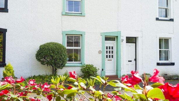 A white building with green trim, a small tree, and red flowers in the foreground. - 3 Bed in Keswick (oc-sz395) (Keswick)