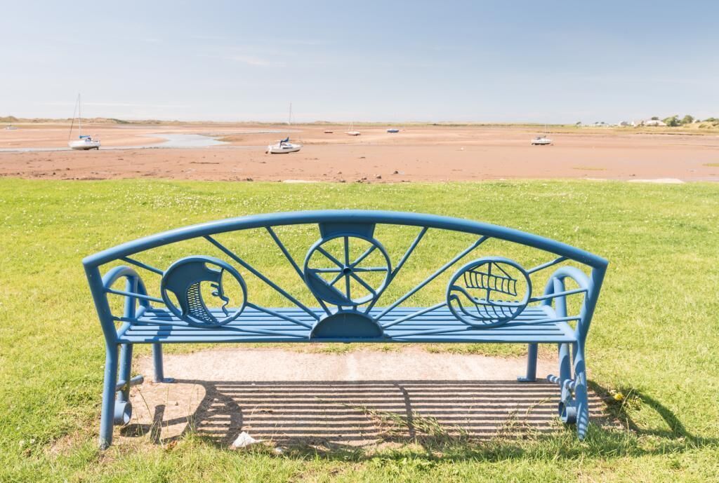 Un banc en métal bleu au design original, installé dans un espace verdoyant, d’où l’on peut admirer une plage et des bateaux au loin.