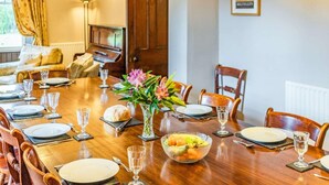 Uma sala de jantar com uma mesa de madeira posta para refeição, cadeiras, um vaso com flores e um piano ao fundo.