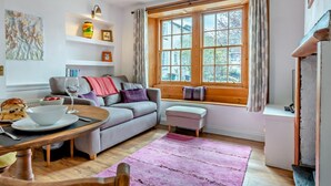 A cozy living room with a sofa, a dining table, a pink rug, and a large window with curtains. - 1 Bed in Keswick (oc-sz472) (Keswick)