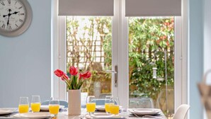 Een eettafel gedekt met borden, glazen en een vaas rode tulpen, met een klok aan de muur en uitzicht op groen buiten.