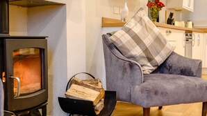 A cozy living room with a fireplace, a grey armchair, and a stack of firewood. - 2 Bed in Keswick (oc-sz036) (Keswick)