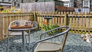 A stone building with a wooden fence, a small table with chairs, and a garden area with plants and flowers. - 2 Bed in Keswick (oc-sz026) (Keswick)