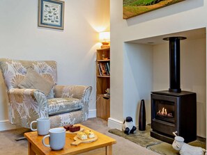 Ein gemütliches Wohnzimmer mit einem gemusterten Sessel, einem Holztisch mit Tassen und Snacks, einem Bücherregal und einem Kaminofen.