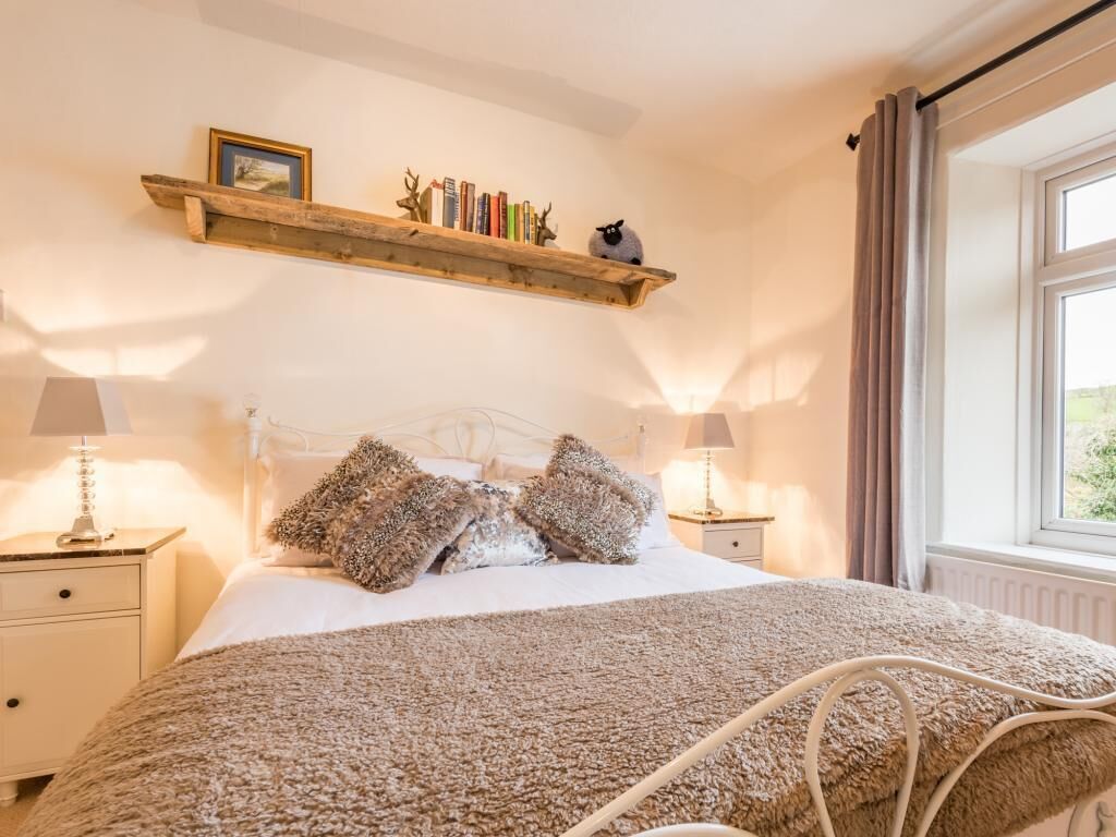 A bedroom with a bed, two bedside tables, a wooden shelf with books and a sheep decoration, and a window with curtains.