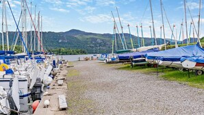 Uma marina com barcos atracados, um caminho de cascalho e montanhas ao fundo.