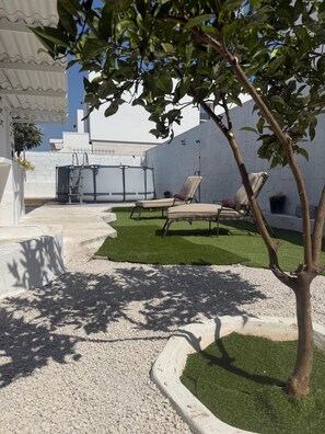 Eine Terrasse mit Kunstgr Rasen, zwei Liegestühlen, einem kleinen Baum und einem Wasserspiel.
