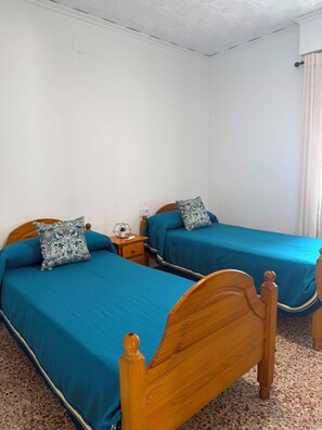 A room with two wooden beds with blue bedding, a small bedside table with a lamp, and a wall socket.