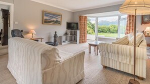 A living room with a sofa, armchair, TV, and a large window with a view of greenery. - 3 Bed in Keswick (oc-sz528) (Portinscale)