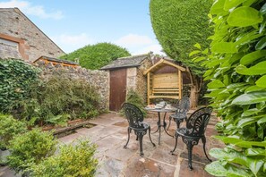 A stone patio with a wooden table and chairs, surrounded by lush greenery and a stone wall. - 2 Bed in Penrith (oc-sz276) (Stainton)