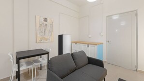 A small, modern room with a gray sofa, a dining table with chairs, a kitchenette, and a white door. - Barrio Sanctuary - Nova (San Juan)