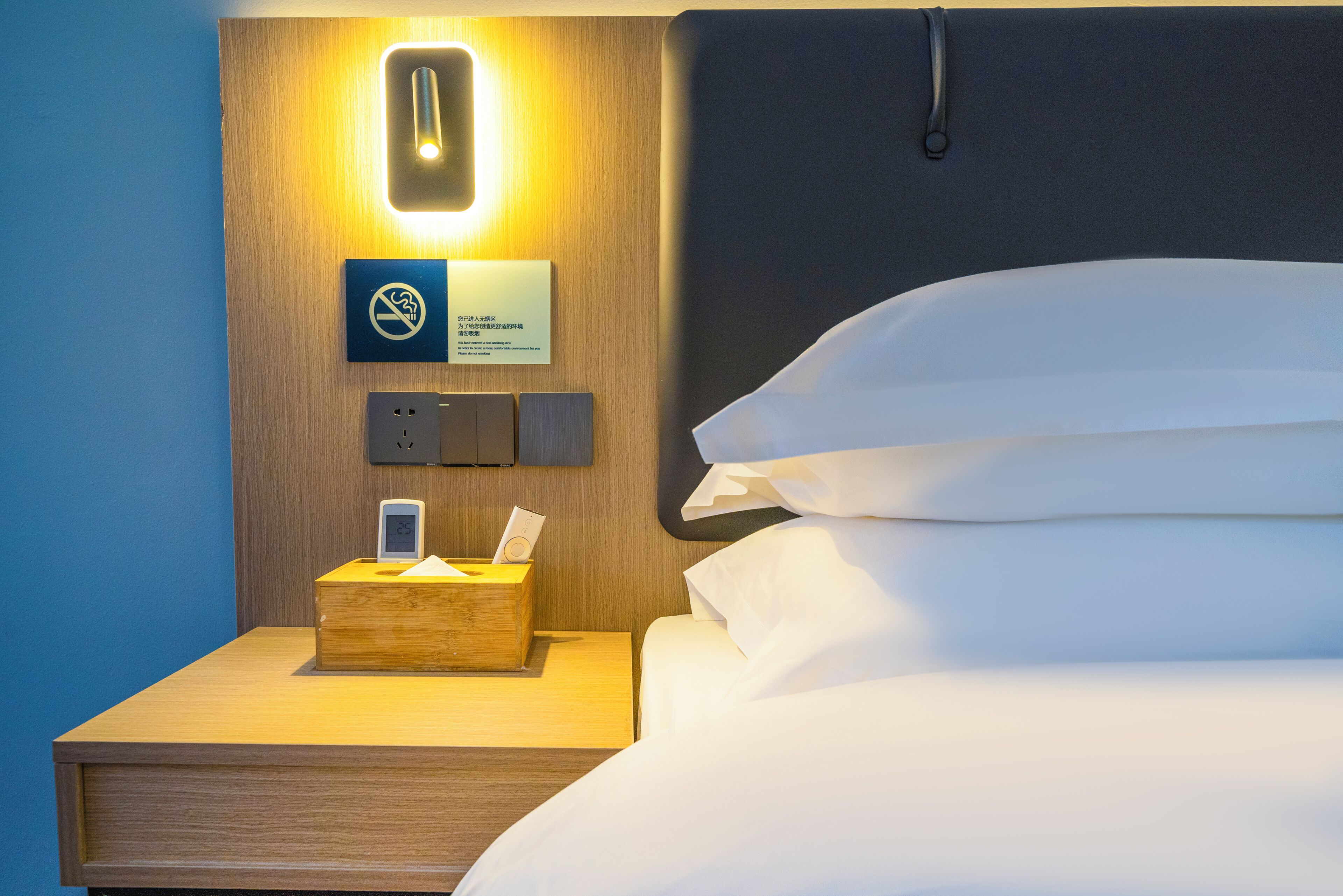Et hotelværelse med en pænt redt seng, et sengebord med en lampe, en vægmonteret lampe og et skilt med rygning forbudt.