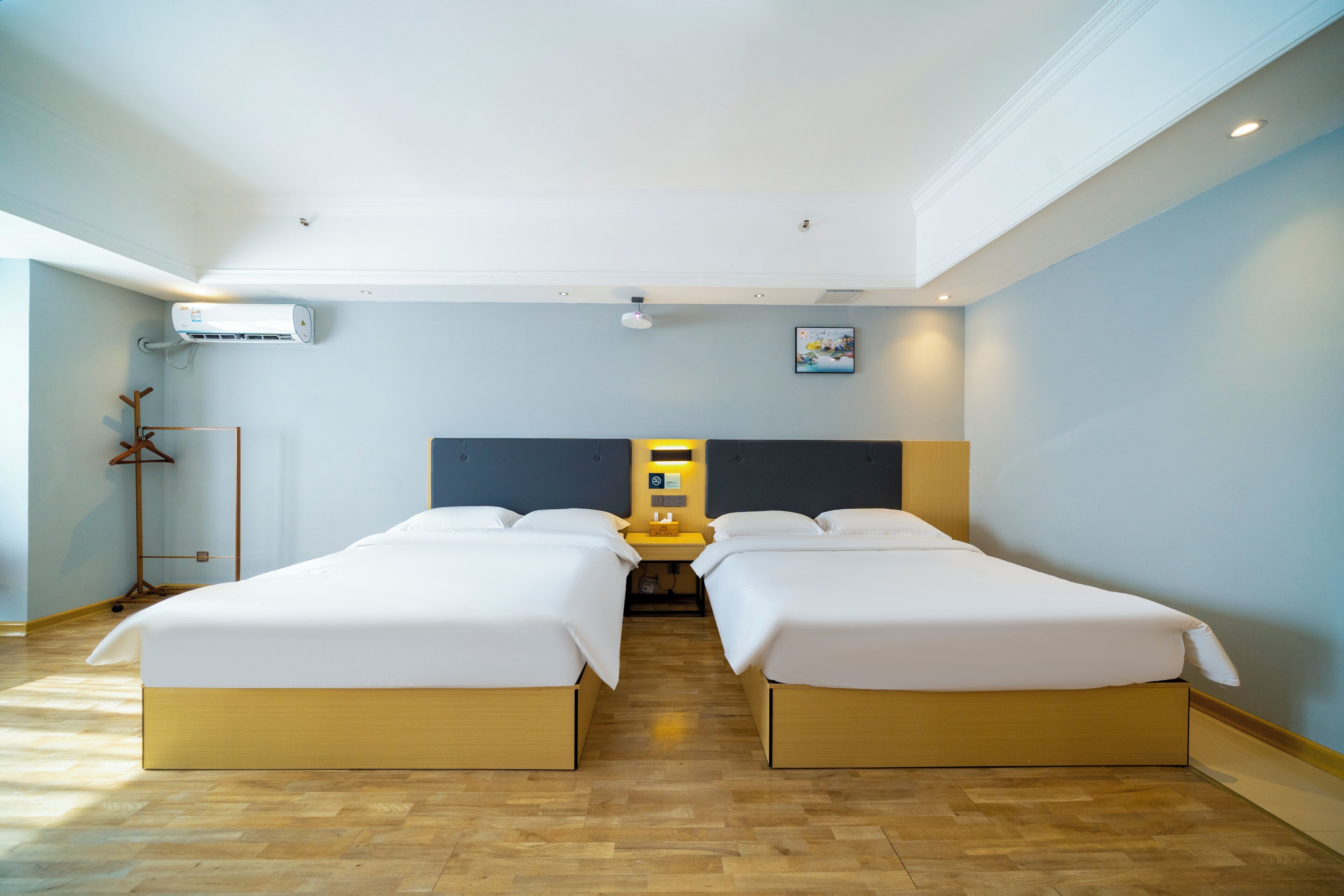 A hotel room with two beds, a bedside table, an air conditioning unit, and a floor lamp.