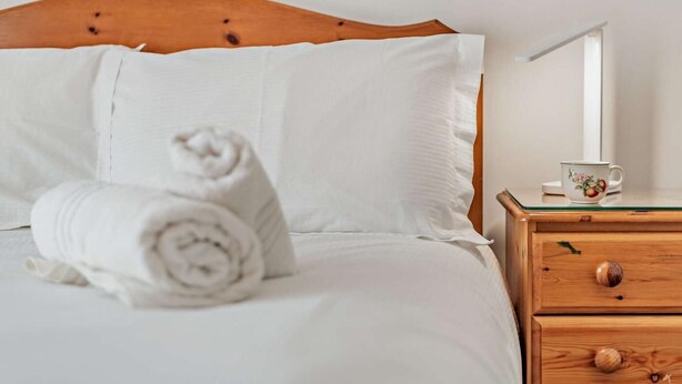 Een keurig opgemaakt bed met witte lakens, een houten hoofdbord, een opgerolde handdoek, een nachtkastje met een kopje en een lamp.