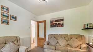 Een woonkamer met een bank en fauteuil, een deur naar een andere kamer, en een wand met ingekaderde foto's.