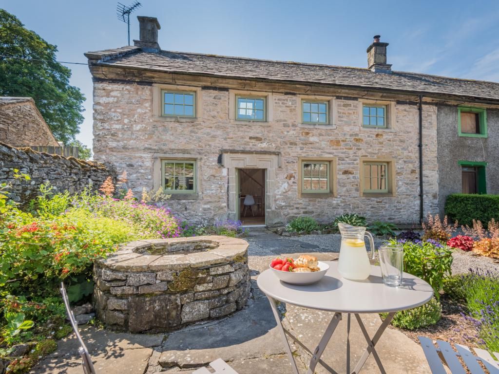 A stone building with a garden in front, a stone bench, and a table with a pitcher and glass.