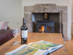 A wooden table with a wine bottle and two glasses, an open magazine, and a fireplace in the background. - 3 Bed in Penrith (oc-sz390) (Shap)