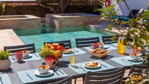 Uma área de refeições junto à piscina, com uma mesa posta para o pequeno-almoço, rodeada de cadeiras e uma piscina ao fundo.