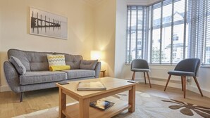 Een woonkamer met een bank, een salontafel, twee stoelen, een lamp en een raam met jaloezieën.