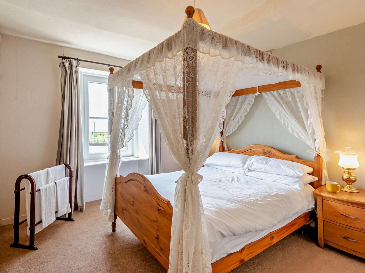 A four-poster canopy bed with a wooden frame and white bedding, a bedside table with a lamp, and a window with curtains.