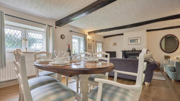 Un coin repas avec une table ronde, des chaises blanches dotées de coussins à carreaux, une cheminée et un canapé, le tout dans une pièce au plancher de bois et aux poutres apparentes.