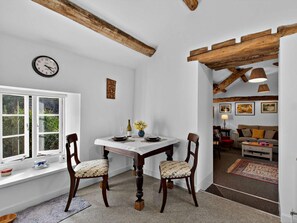 Zona de menjador amb taula blanca, dues cadires, un rellotge i una finestra que mostra vistes a la vegetació.