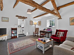 A spacious living room with exposed wooden beams, a television, bookshelves, a red armchair, and a patterned rug. - 1 Bed in Carnforth (oc-sz527) (Barbon)