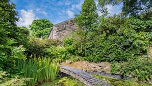 Un ponte in pietra sopra uno stagno, con vegetazione circostante e un edificio in pietra sullo sfondo.