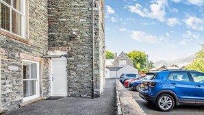 Un edificio de piedra con una puerta y ventanas blancas, un estacionamiento con coches, y cielo despejado.