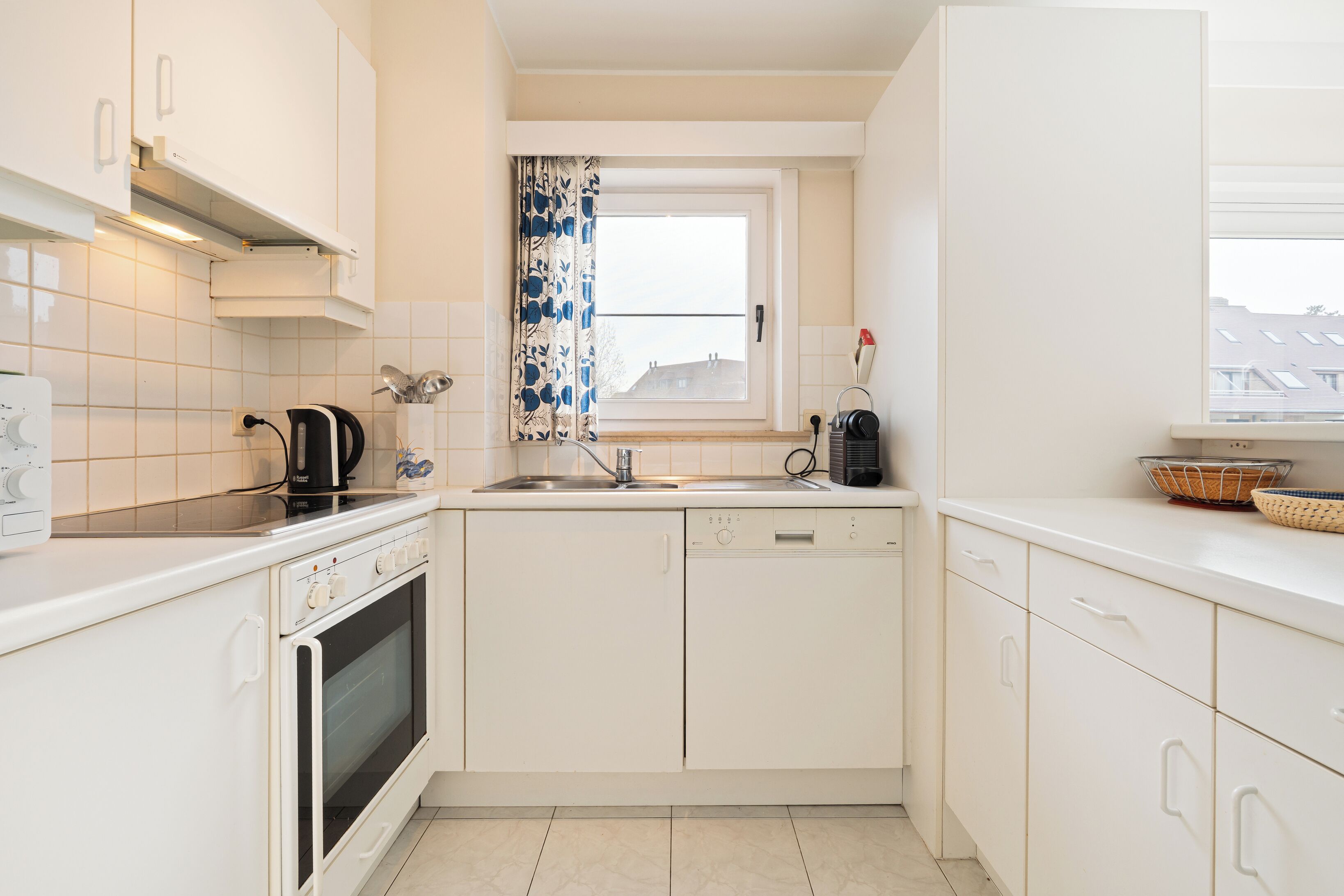 Une cuisine compacte avec des meubles blancs, un four encastré, un lave-vaisselle et une fenêtre avec un rideau.