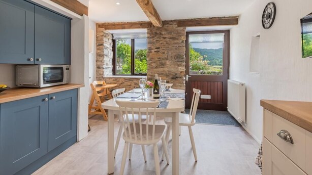 A kitchen with a stone wall, wooden beams, a dining table with chairs, and a view of the outdoors. - 2 Bed in Ulverston (oc-sz591) (Haverthwaite)