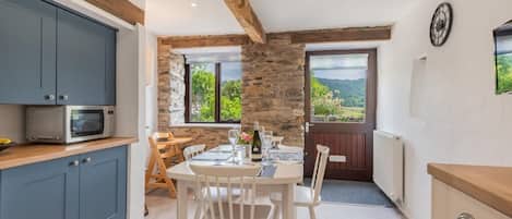 A kitchen with a stone wall, wooden beams, a dining table with chairs, and a view of the outdoors.