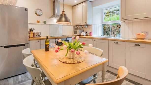 A modern kitchen with white cabinets, a stainless steel refrigerator, a wooden dining table with a vase of flowers, and a hanging pendant light. - 3 Bed in Penrith (oc-sz184) (Glenridding)