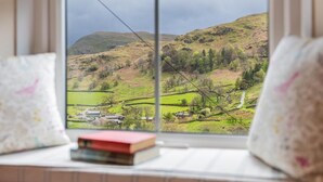 A window with a view of a green landscape, a book on the windowsill, and a pillow. - 3 Bed in Penrith (oc-sz184) (Glenridding)