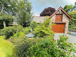 A stone building with a wooden door and a small garden area with various plants and a fountain. - 4 Bed in Holmrook (oc-sz293) (Irton with Santon)
