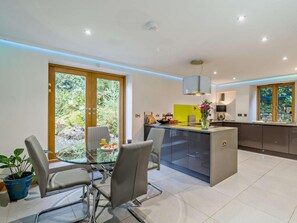 A modern kitchen with a central island, a dining area with a glass table, and a view of greenery through large sliding doors. - 4 Bed in Holmrook (oc-sz293) (Irton with Santon)