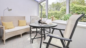 A sunlit patio with a wicker sofa, a round table, and a chair, overlooking a garden. - 2 Bed in Keswick (oc-sz188) (Keswick)