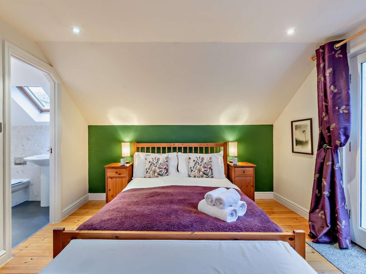 Een slaapkamer met schuine wanden, een houten bed, een groene accentmuur, twee nachtkastjes met lampen, een raam met uitzicht, en een deur met paarse gordijnen.