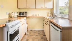 A compact kitchen with a microwave, oven, and sink, wooden cabinets, and a window providing a view. - 1 Bed in Kirkby-in-furness (oc-sz463) (Kirkby Ireleth)