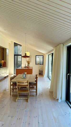 A spacious dining area with a wooden table and chairs, a kitchen in the background, and large windows allowing natural light. - Beautiful cottage with a simple modern aesthetic (Vejby Strand)