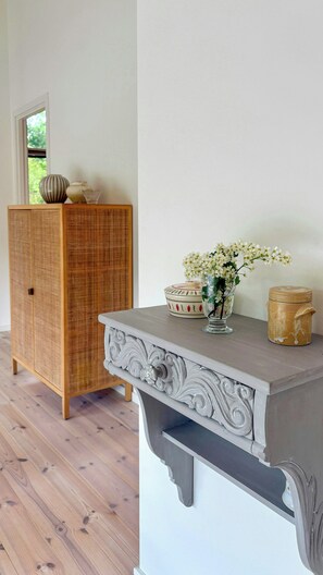 A room with a wooden cabinet, a decorative console table with a vase of flowers, and a window with a view of greenery. - Beautiful cottage with a simple modern aesthetic (Vejby Strand)